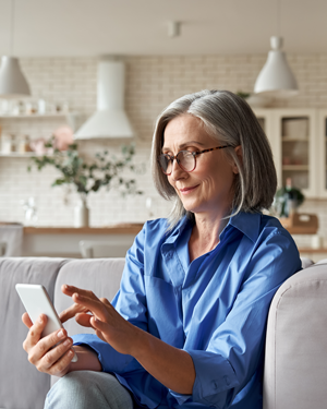 Elger Dental | Smiling older woman with gray hair using a smartphone while sitting on a couch at home